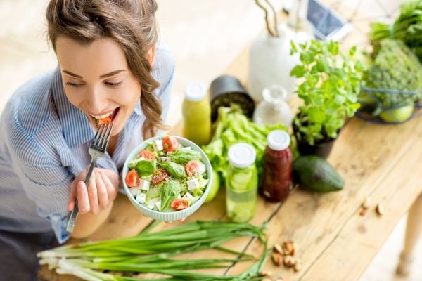  Mulher jovem e feliz comendo salada saudável, sentado à mesa com ingredientes frescos verdes dentro de casa