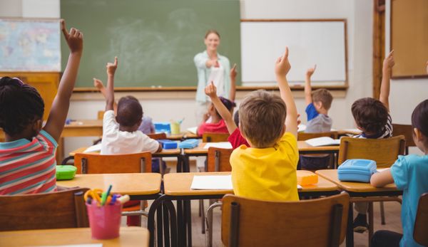 Crianças na sala de aula levantando o dedo para responder a pergunta da professora