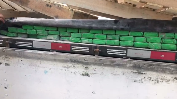 A imagem mostra vários pacotes de maconha embalados em um plástico verde escondidos por baixo de uma lona no compartimento de carga de um caminhão
