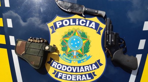 A imagem mostra o revólver e as munições junto de um coldre de arma e uma faca em cima de uma superfície com o emblema da Polícia Rodoviária Federal ao fundo.
