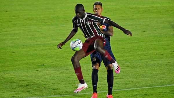 A imagem mostra um jogador do Fluminense dominando a bola, marcado de perto por outro jogador do Bragantino