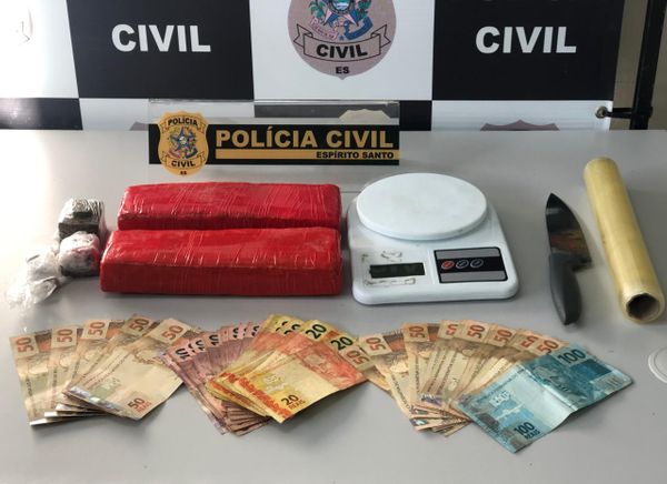 A imagem mostra várias notas de real sobre uma mesa com dois papelotes de maconha, uma balança, uma faca, um rolo de papel manteiga e um distintivo da Polícia Civil.