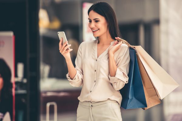 Mulher segurando sacolas e olhando o celular; compras online