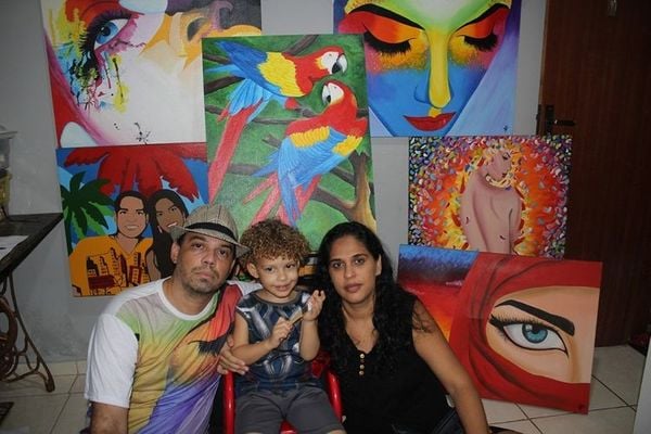 O professor e artista plástico Gustavo Gomes posa com esposa e filho em frente a suas obras. O quadro do centro está sendo rifado para manter as contas em dia