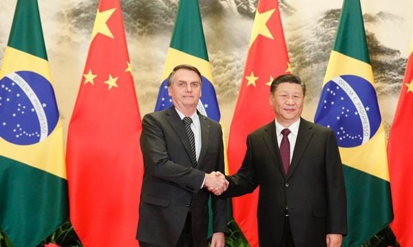 Xi Jinping e Bolsonaro apertam as mãos em frente a bandeiras chinesas e brasileiras.