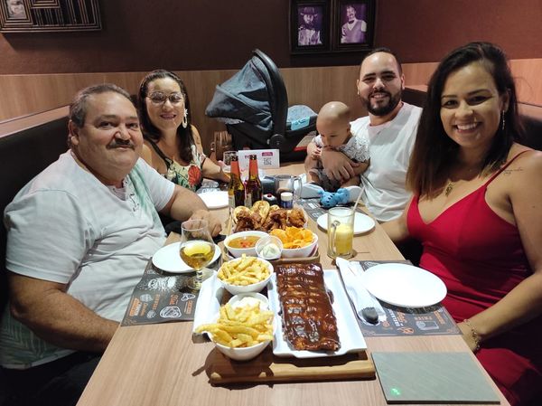 Pai, mãe, esposa e filho de Lucas, além dele próprio, sentados ao redor de uma mesa de comida e posando para a foto
