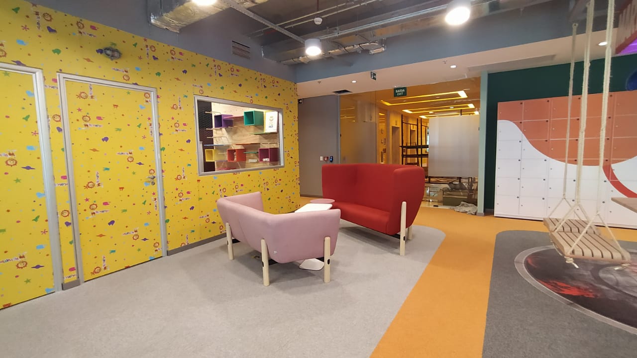 Ambiente corporativo com paredes amarelas, dois sofás assimétricos, um vermelho e outro rosa no centro. No canto direito há um balanço. 