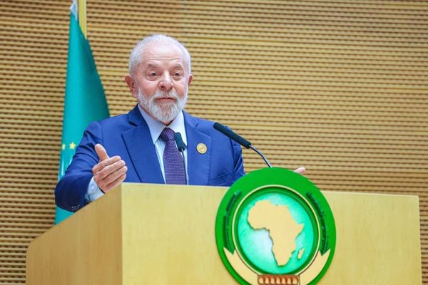 Lula é um homem idoso, branco, com cabelos e barba curtos e brancos; ele veste terno azul-escuro, camisa azul-bebê e gravata roxa; ele discursa em um púlpito O presidente Lula discursa em cúpula da União Africana, na Etiópia