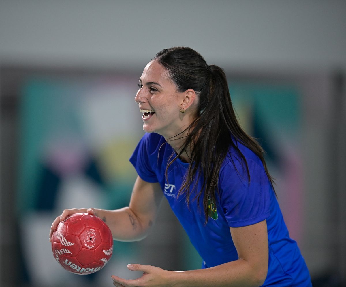 Atleta de uniforme azul, segurando uma bola de handebol vermelha, em treino da seleção brasileira feminina