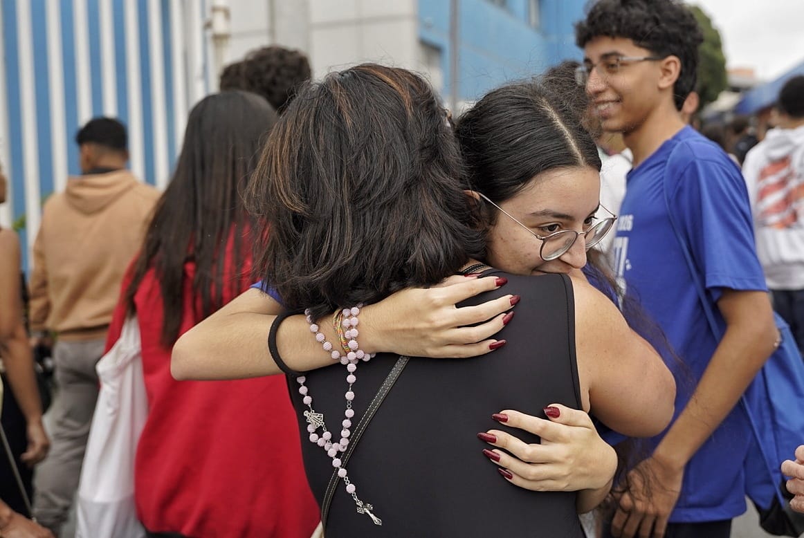 Duas mulheres se abraçando. Uma delas tem um terço em sua mão.